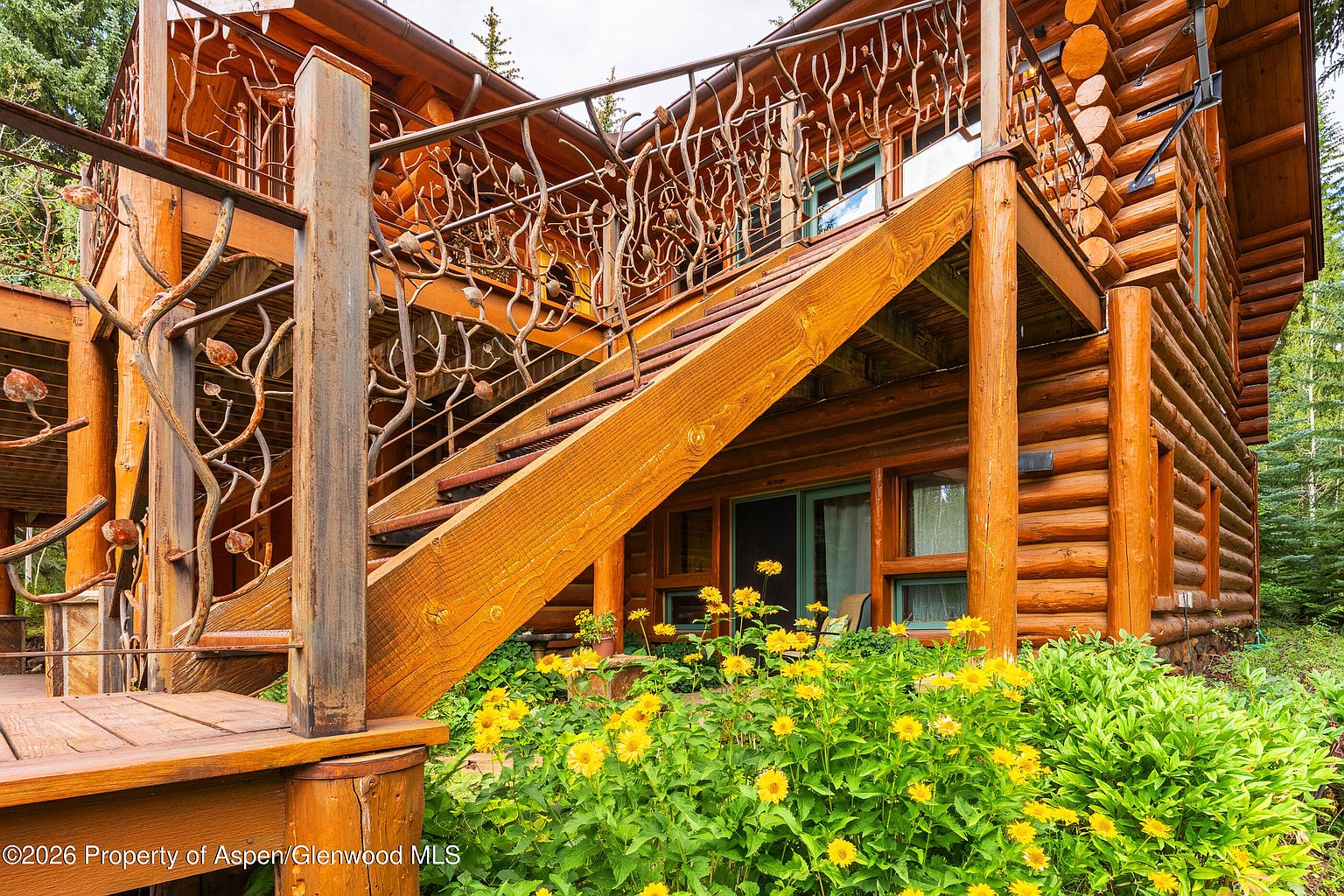 This image captures a rustic log cabin exterior, focusing on a prominent wooden staircase leading to an upper-level deck. The deck features unique, organic-shaped metal railings that mimic the appearance of twisted branches, adding a distinct artistic flair to the structure. Below the deck, a lush garden filled with vibrant yellow flowers frames the lower level, which includes a glass door and windows, creating a warm and inviting mountain retreat aesthetic.