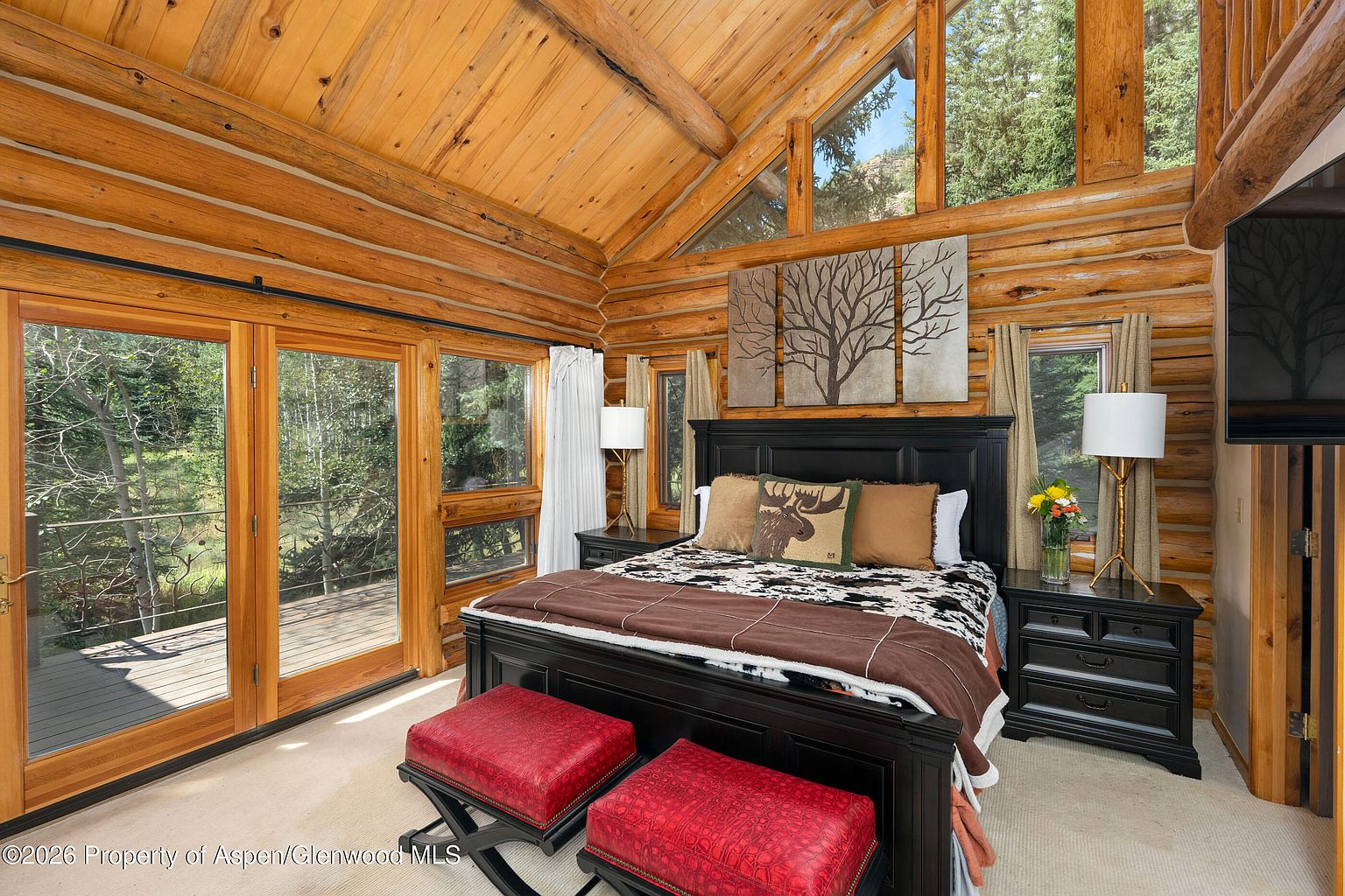This rustic primary bedroom features exposed log walls and a vaulted wood-paneled ceiling, creating a warm, cabin-inspired atmosphere. A large black bed frame serves as the centerpiece, complemented by two red upholstered ottomans at the foot and a set of three tree-themed wall art pieces above. Large windows and glass doors provide abundant natural light and views of the surrounding forest, while two bedside lamps add a soft, inviting glow to the space.