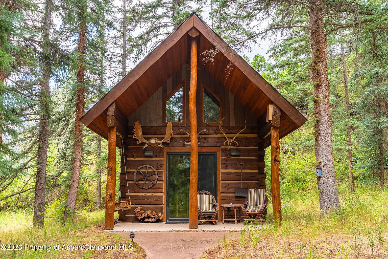 This charming rustic log cabin is nestled within a dense forest, featuring a prominent A-frame roofline and a covered front porch. The exterior is constructed from dark, horizontal logs and is decorated with mounted antlers and a decorative wagon wheel. A small seating area with two chairs and a hanging swing provides a cozy space to enjoy the surrounding natural landscape.