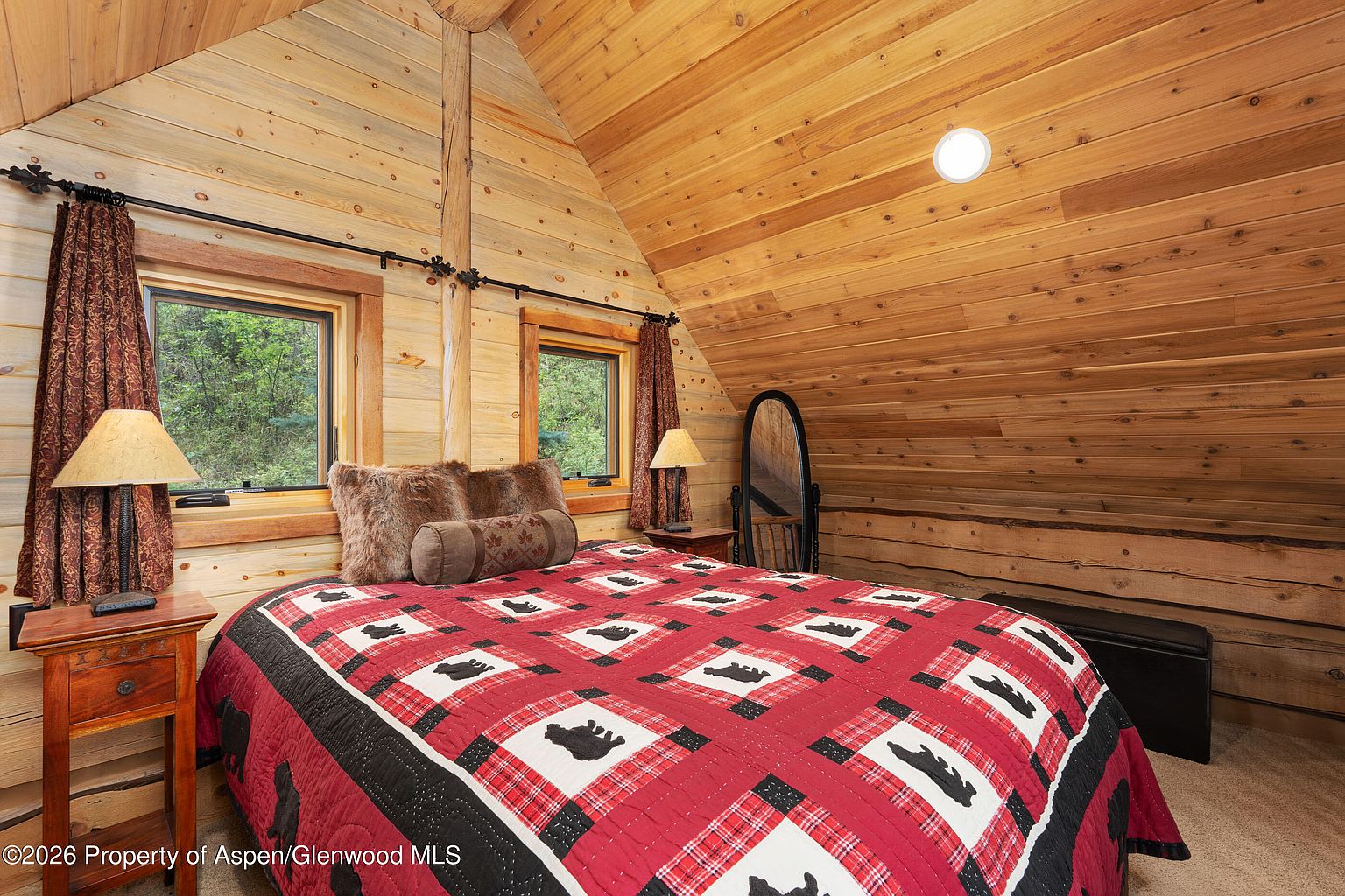 This cozy bedroom features a rustic log cabin aesthetic with warm wood-paneled walls and a vaulted ceiling. The room is centered around a bed dressed in a red and black plaid quilt with bear motifs, flanked by two wooden nightstands with matching lamps. Two windows with dark patterned curtains provide natural light, while a full-length mirror stands in the corner, creating a comfortable and inviting mountain retreat atmosphere.