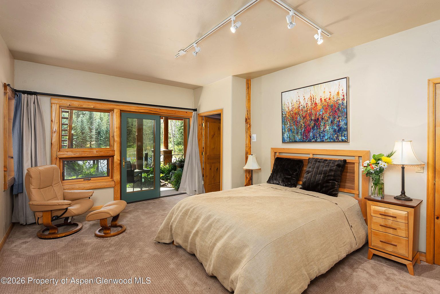 This cozy bedroom features a neutral-toned bed with dark accent pillows, positioned beneath a vibrant, abstract floral painting. To the left, a leather recliner with an ottoman sits near large windows and a glass door that opens to a lush, wooded outdoor view. The room is finished with light wood trim, a matching nightstand with a lamp, and track lighting mounted on the ceiling.