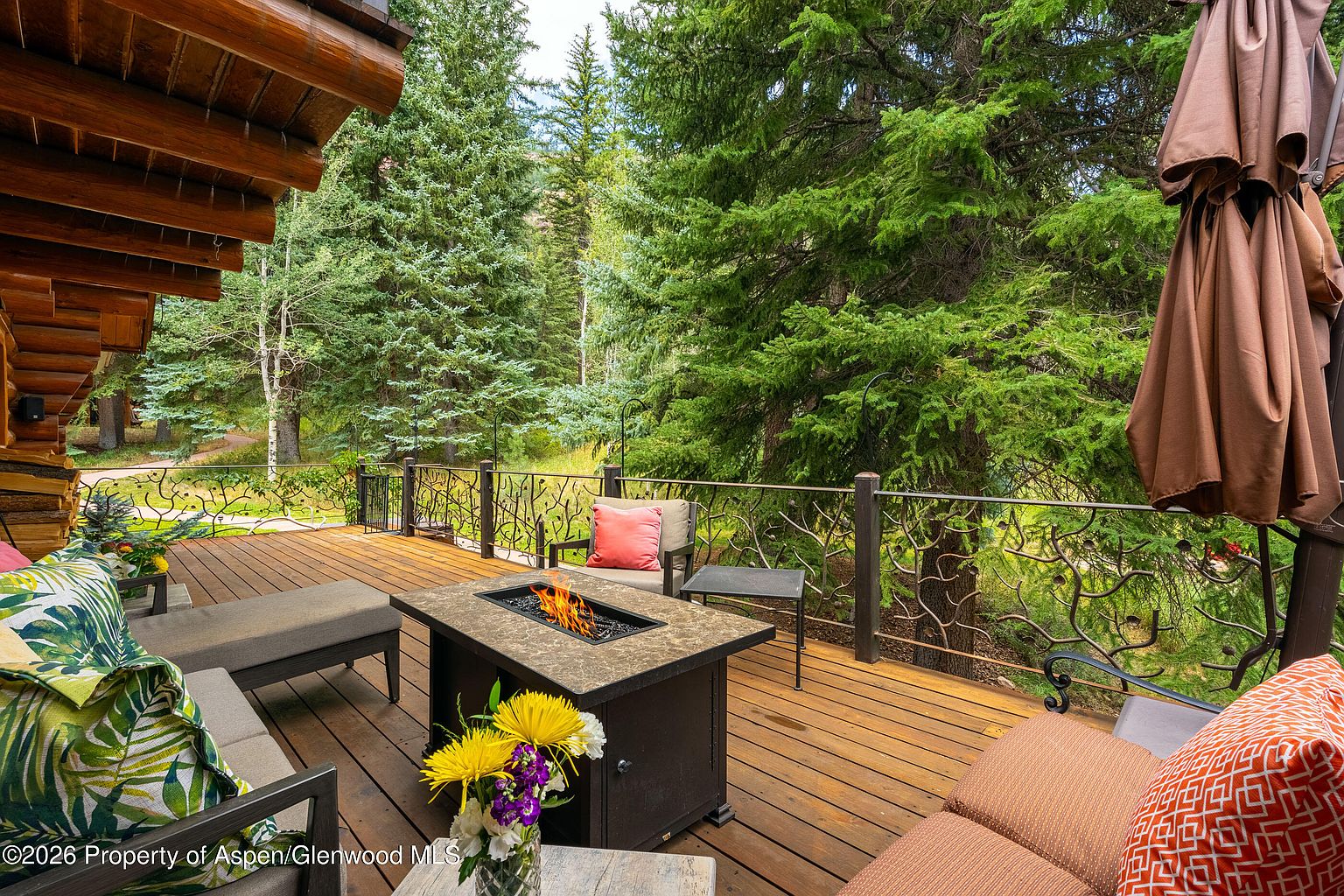 This elevated wooden deck offers a serene outdoor living space nestled among mature evergreen trees. The area is furnished with comfortable lounge seating and a central fire table, creating an inviting atmosphere for relaxation. A decorative metal railing with a branch-like design provides safety while maintaining an unobstructed view of the lush, wooded surroundings.