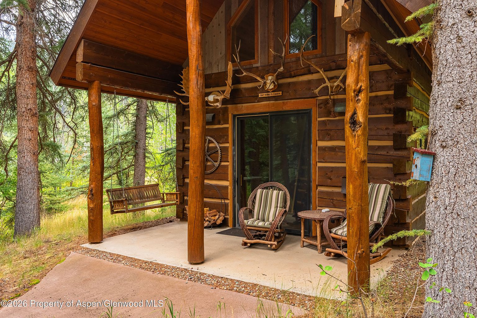 This rustic log cabin features a covered front porch area complete with a wooden swing, two rocking chairs, and a small side table. The exterior is constructed from stacked logs, adorned with decorative antlers and a sign above the sliding glass door, creating a cozy, woodland retreat atmosphere. The porch overlooks a lush, forested landscape, emphasizing a peaceful and secluded mountain setting.