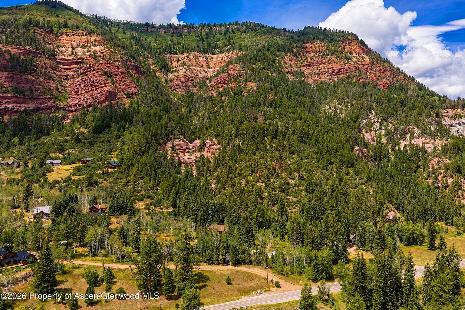 This wide-angle aerial perspective captures a sprawling mountain landscape featuring a dense forest of evergreen trees set against dramatic, layered red rock cliffs. Several residential homes are nestled into the hillside, connected by winding dirt roads that lead down to a paved highway in the foreground. The scene conveys a sense of seclusion and natural beauty, highlighting the property's integration into the rugged Colorado terrain.