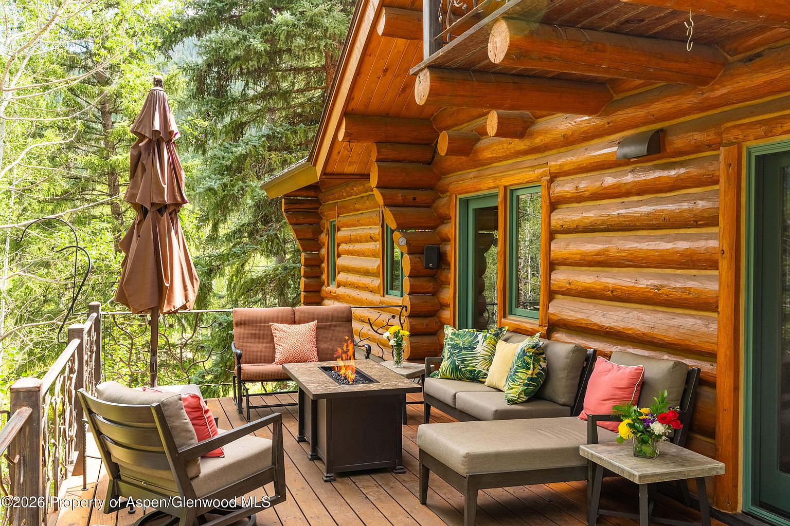 This inviting wooden deck features a rustic log cabin exterior and is furnished with comfortable outdoor seating, including a sofa and armchairs arranged around a central fire pit. The space is surrounded by lush forest greenery, creating a serene and private atmosphere perfect for relaxation. The perspective captures the cozy, elevated outdoor living area from a slightly elevated angle, highlighting the blend of natural wood textures and comfortable modern furnishings.