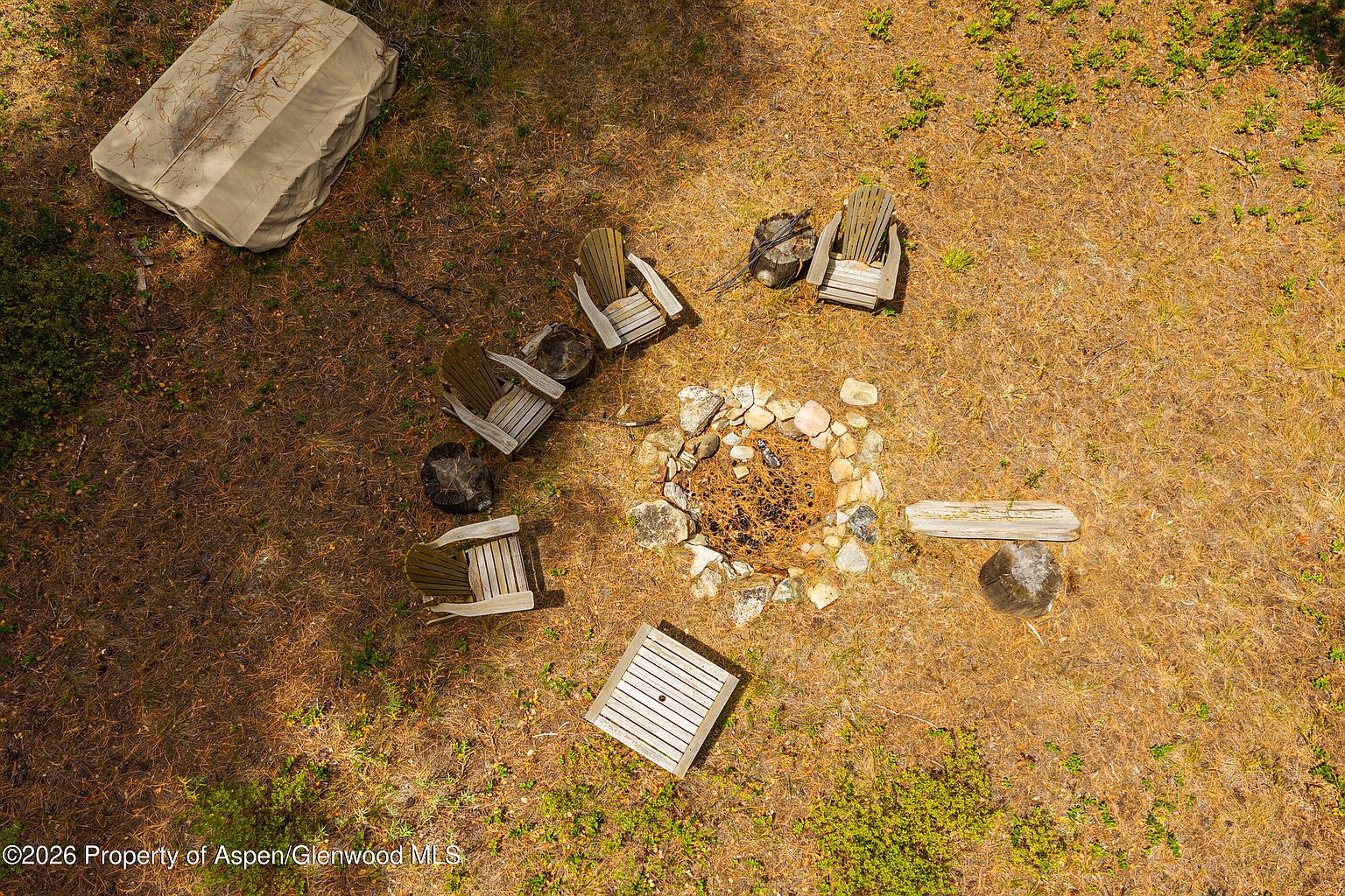 This high-angle aerial view captures a rustic outdoor fire pit area nestled within a natural, wooded landscape. The scene features a circular stone-lined fire pit surrounded by several wooden Adirondack chairs, a small side table, and natural log seating. The ground is covered in a layer of pine needles, creating a serene and secluded atmosphere perfect for outdoor gatherings.