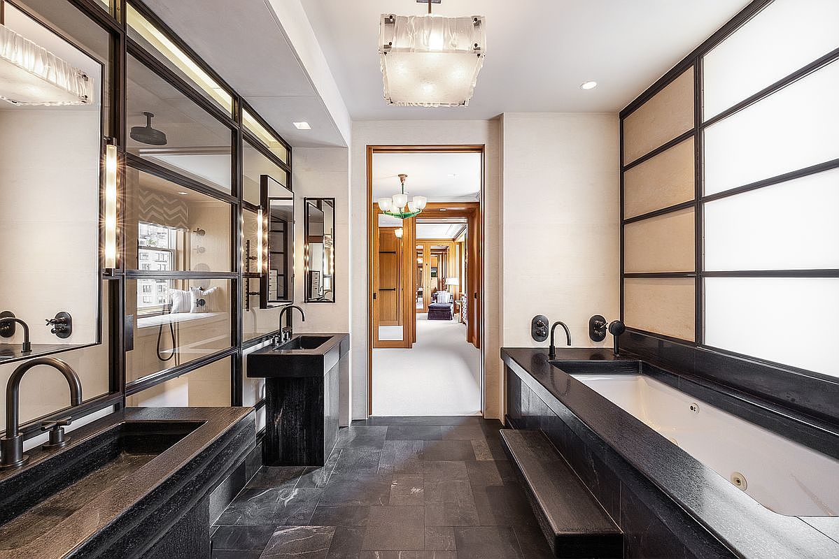 This luxurious primary bathroom features a sophisticated, dark-toned aesthetic with stone surfaces and a dramatic, symmetrical layout. A deep soaking tub is positioned against a wall with frosted glass panels, while dual vanity sinks are integrated into dark stone counters on either side. The perspective looks through the center of the room toward an open doorway that reveals a bright, carpeted hallway and additional living spaces beyond.