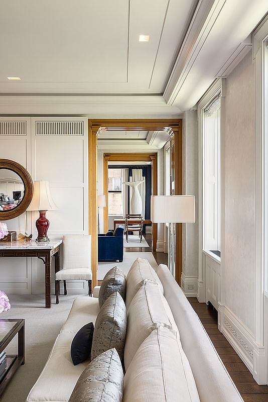 This elegant living room features a neutral color palette with a long, light-toned sofa positioned in the foreground, accented by textured throw pillows. A console table with a decorative lamp and a round mirror sits against a paneled wall, while a wide doorway leads into a series of interconnected rooms, creating a sense of depth and architectural flow. The space is characterized by sophisticated millwork, high ceilings with recessed lighting, and a bright, airy atmosphere.
