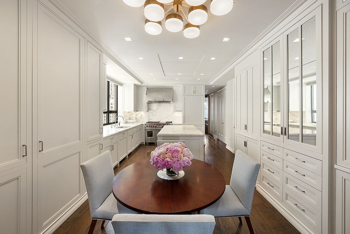 This elegant, high-end kitchen features a central dining area with a round wooden table and upholstered chairs, accented by a vibrant floral centerpiece. The space is characterized by custom white cabinetry, a central island with a stone countertop, and a professional-grade range with a stainless steel hood. The perspective is a straight-on, symmetrical shot looking through the kitchen toward the back, highlighting the sophisticated lighting fixtures and rich dark wood flooring.