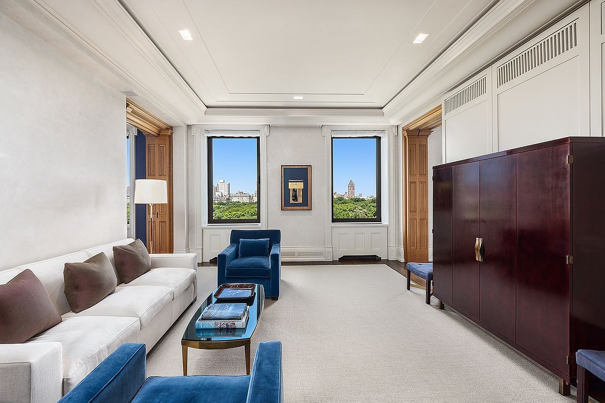 This elegant living room features a sophisticated color palette with a white sofa, deep blue velvet armchairs, and a dark wood cabinet. Two large windows frame a scenic view of a park and city skyline, allowing ample natural light to fill the space. The room is finished with crown molding, light-toned walls, and a neutral area rug, creating a refined and airy atmosphere.