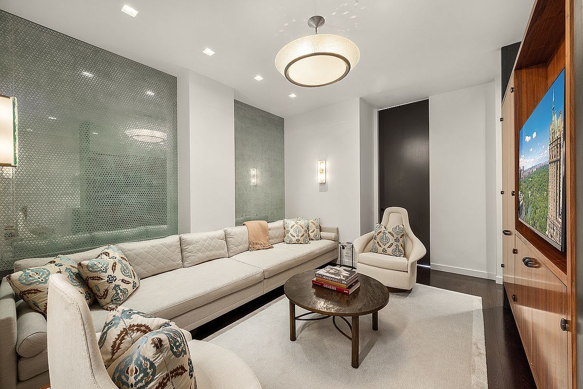 This sophisticated living room features a long, neutral-toned sectional sofa accented with patterned throw pillows, centered around a round wooden coffee table. The space is defined by textured, light-colored wall panels and a modern, circular pendant light fixture that provides a soft glow. A large television is mounted on a custom wood-paneled wall unit, creating a sleek and contemporary entertainment area.