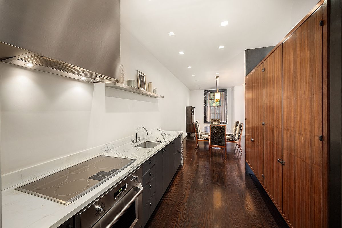 This sleek, galley-style kitchen features dark cabinetry paired with light-toned stone countertops and a stainless steel range hood. A long wall of warm-toned wood cabinetry provides extensive storage, while the perspective leads the eye toward a formal dining area at the end of the space. The room is finished with dark wood flooring and recessed lighting, creating a sophisticated and streamlined aesthetic.