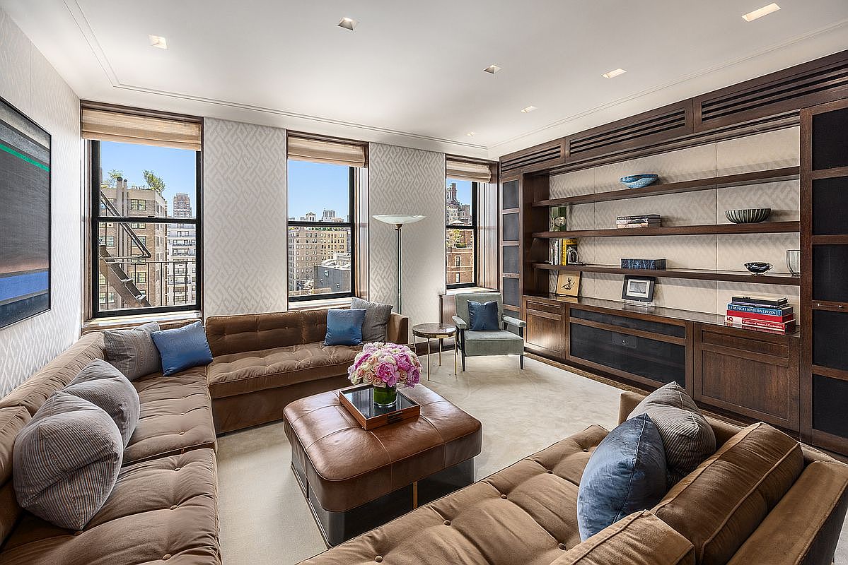 This sophisticated living room features a large, plush brown sectional sofa arranged around a matching leather ottoman, creating a cozy and inviting seating area. A wall of custom dark wood shelving provides ample display space, while large windows allow natural light to fill the room and offer views of the city skyline. The space is accented by a patterned wallpaper, a soft area rug, and a single armchair, resulting in a refined and comfortable atmosphere.