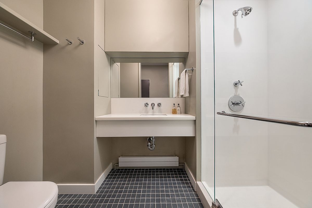 This modern bathroom features a floating vanity with a white countertop and a rectangular sink, set against a neutral-toned wall. To the right, a glass-enclosed shower stall with a chrome fixture adds a sleek, contemporary touch, while the floor is finished in dark, small-format square tiles. The space is efficiently designed with a wall-mounted mirror and a towel rack, creating a clean and functional aesthetic.