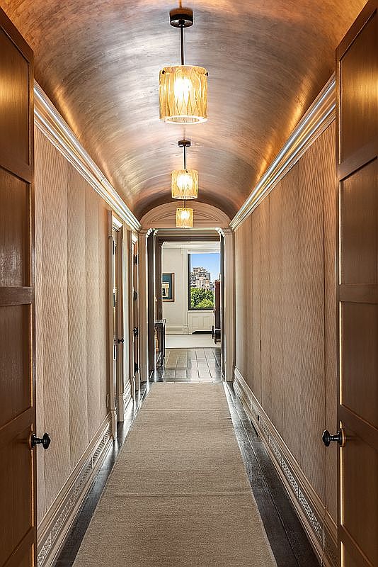 This elegant, long hallway features a dramatic barrel-vaulted ceiling with a metallic finish, illuminated by a series of three matching pendant lights. The walls are covered in a textured, neutral-toned wallcovering, and a long runner rug centers the dark wood flooring. The perspective looks down the corridor toward a bright, sunlit room with a window view, creating a sense of depth and sophisticated transition.