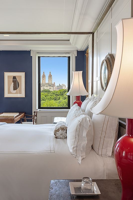 This elegant primary bedroom features a sophisticated navy blue accent wall and a four-poster bed dressed in crisp white linens. A large window provides a stunning, direct view of a lush green park and iconic city architecture, while a vibrant red lamp adds a bold pop of color to the bedside table. The room exudes a refined, classic aesthetic with high-end finishes and a serene, luxurious atmosphere.