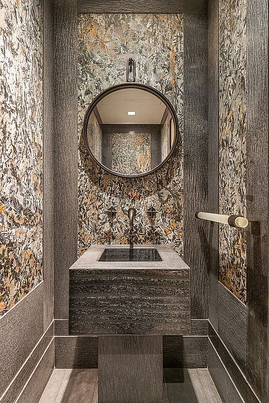 This sophisticated powder room features a striking, textured stone sink pedestal and a matching wall-mounted faucet. The space is framed by dark, vertical-grain wood paneling and dramatic, patterned stone wall cladding that creates a moody, high-end aesthetic. A circular mirror is centered above the sink, reflecting the intricate patterns of the wall behind it.