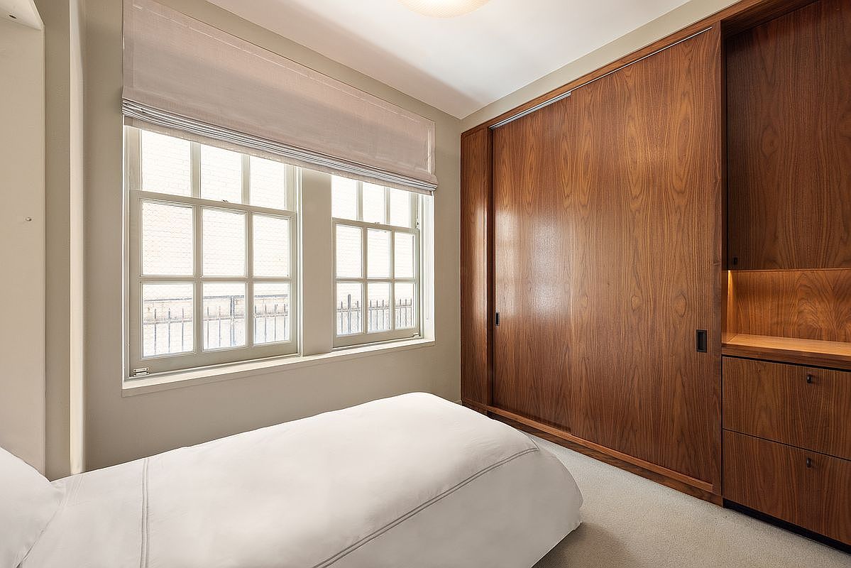 This compact bedroom features a minimalist aesthetic with a single bed dressed in crisp white linens. A large, custom-built wooden storage unit with sliding doors and integrated shelving occupies the entire side wall, providing ample organization space. The room is illuminated by natural light streaming through two large, multi-pane windows covered by a neutral-toned Roman shade.