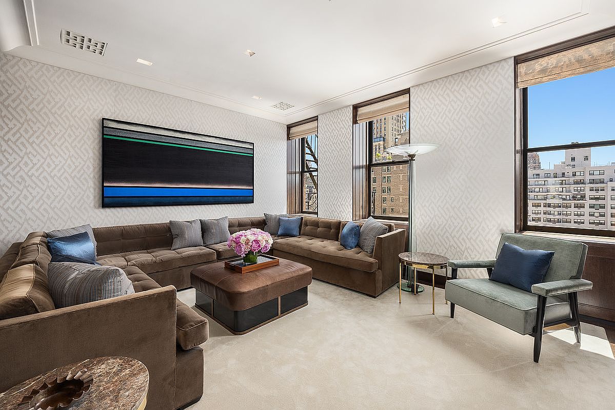This sophisticated living room features a large, L-shaped sectional sofa upholstered in a warm brown fabric, accented with blue throw pillows and a central leather ottoman. The walls are adorned with a subtle, textured geometric wallpaper and a prominent piece of modern abstract art, while tall windows provide ample natural light and city views. A plush armchair and a small side table complete the seating arrangement, creating an elegant and inviting atmosphere.