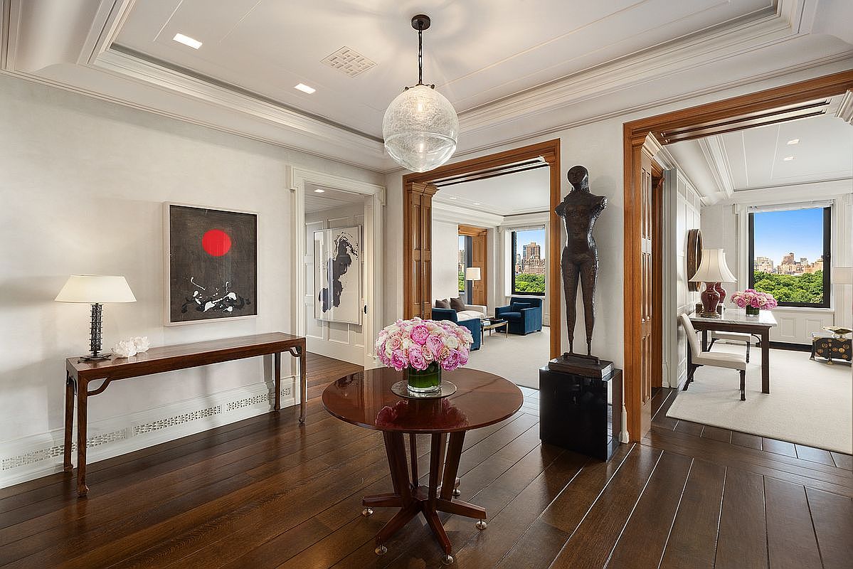 This elegant foyer serves as a central gallery, featuring dark wood flooring and a sophisticated circular pedestal table topped with a vibrant floral arrangement. The space is adorned with curated artwork, a tall sculptural figure, and a distinctive glass pendant light, creating a refined and artistic atmosphere. Through the wide, wood-trimmed doorways, one can glimpse adjacent living and office spaces that offer expansive views of the city skyline and greenery.