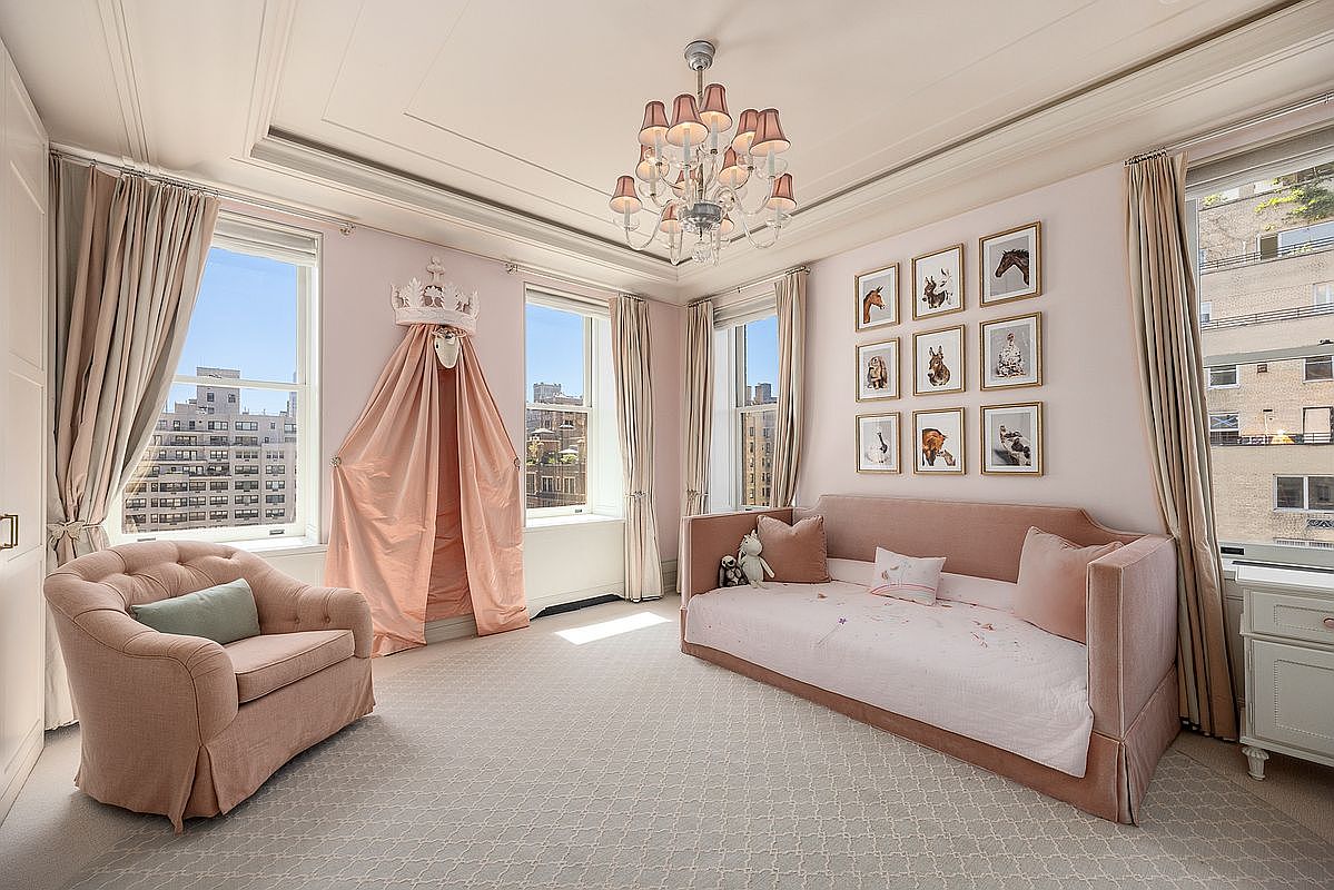 This elegant, light-filled bedroom features a soft pink color palette, a plush daybed, and a matching armchair. A whimsical crown canopy draped in fabric adds a playful touch, while a gallery wall of framed artwork provides a sophisticated focal point. The room is illuminated by a classic crystal chandelier and benefits from abundant natural light streaming through multiple large windows.
