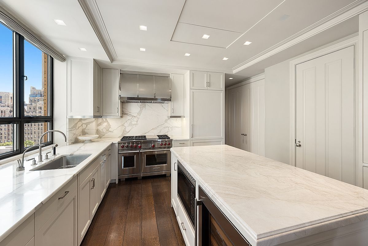 This bright, modern kitchen features white cabinetry, a large central island with a stone countertop, and high-end stainless steel appliances. A large window provides natural light and an urban view, while the space is finished with dark wood flooring and a stone backsplash. The perspective is from the kitchen island looking toward the range, highlighting the clean lines and sophisticated design of the culinary space.