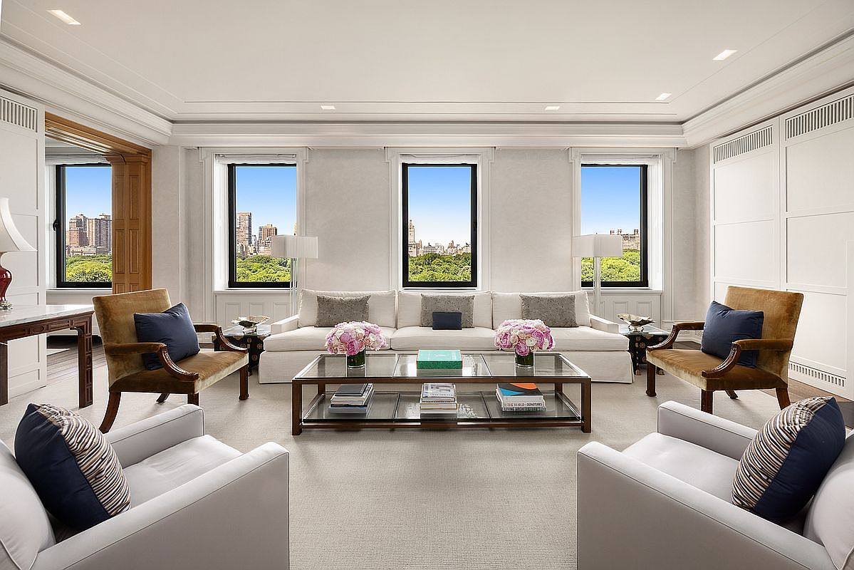 This elegant, symmetrical living room features a large white sofa centered beneath three tall windows that offer expansive views of a park and city skyline. The room is furnished with two plush armchairs in the foreground and two velvet-upholstered chairs on either side of the sofa, all arranged around a glass-topped coffee table. The neutral color palette, clean lines, and sophisticated decor create a refined and airy atmosphere suitable for formal entertaining.