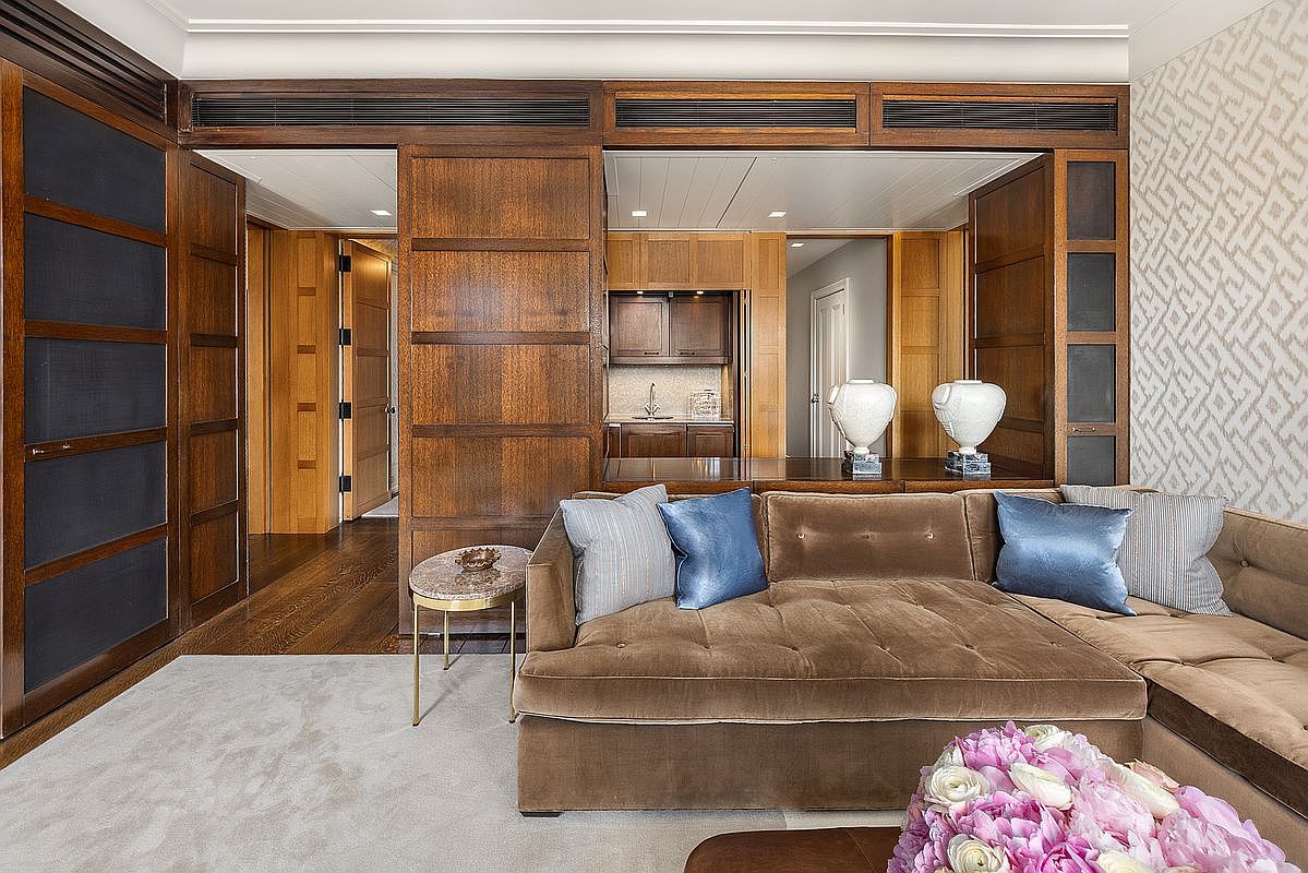 This sophisticated living space features rich wood paneling throughout, creating a warm and traditional atmosphere. A plush, velvet-upholstered sectional sofa sits atop a neutral area rug, accented by blue throw pillows and a bouquet of pink flowers in the foreground. The room opens into a secondary area with a wet bar, showcasing a seamless transition between living and entertaining spaces.