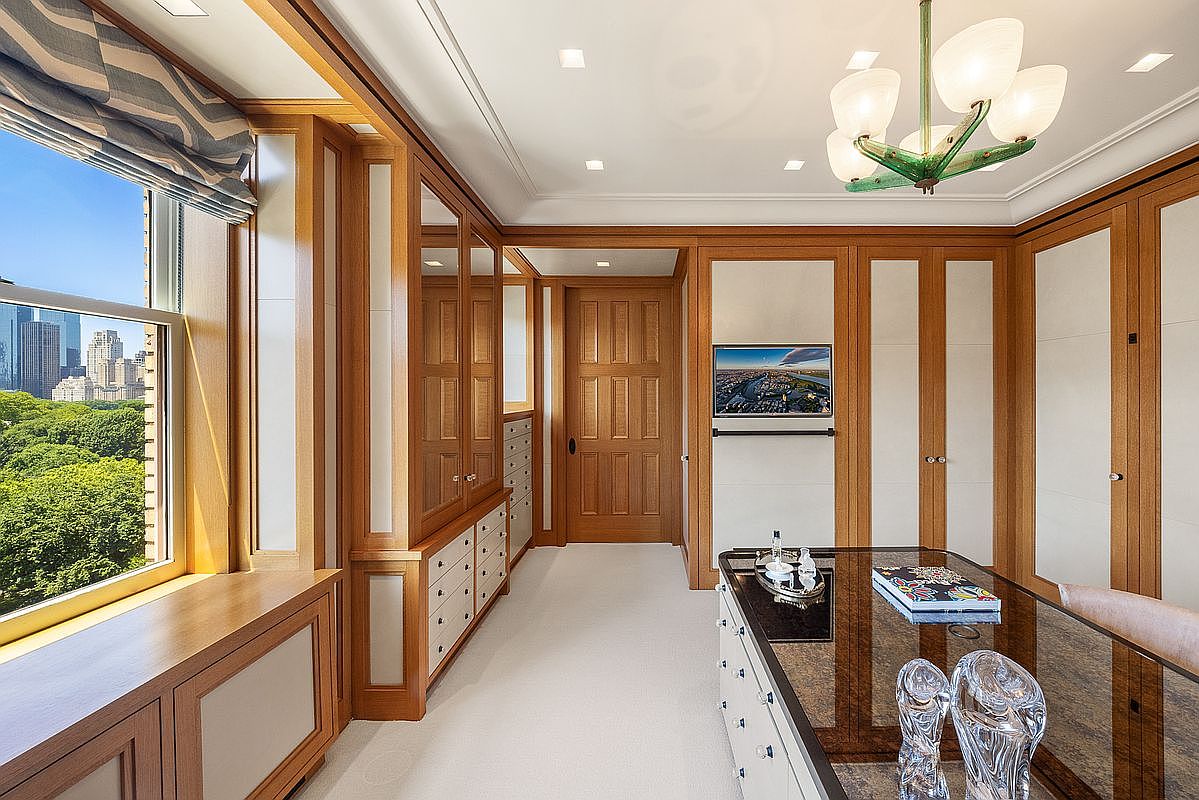 This sophisticated home office features warm wood paneling, built-in cabinetry, and a large desk with a glass top. A window provides a scenic view of a lush green park and city skyline, while a unique, multi-armed light fixture hangs from the ceiling. The room is finished with light-colored carpeting and a central wooden door, creating a professional and refined atmosphere.