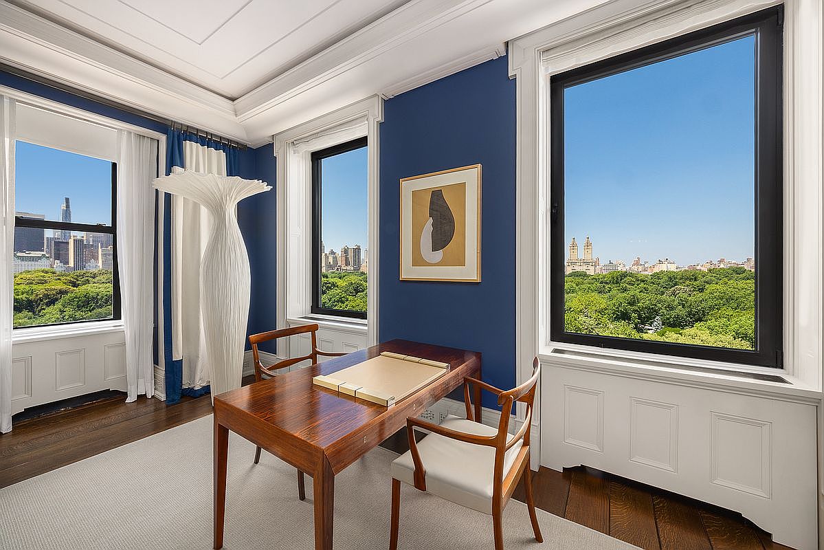 This sophisticated home office features deep blue walls and dark wood flooring, creating a refined and focused atmosphere. The room is anchored by a polished wood desk and two matching chairs, positioned to take advantage of the expansive views of Central Park through large windows. A unique, sculptural floor lamp and a framed piece of abstract art add artistic flair to the space, which is bathed in natural light.