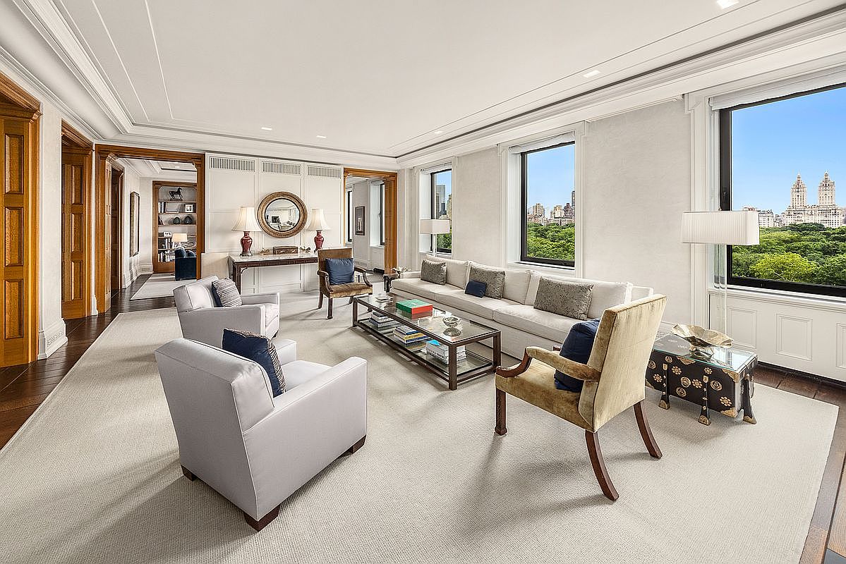 This elegant, sun-drenched living room features a neutral color palette with plush seating, including a long sofa and several armchairs arranged around a glass-topped coffee table. Large windows provide expansive views of a lush green park and city skyline, filling the space with natural light. The room is accented by classic architectural molding, dark wood flooring, and a console table topped with decorative lamps and a circular mirror.