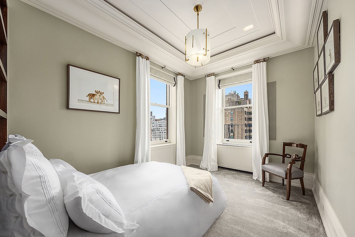 This serene bedroom features a neutral color palette with soft sage walls and plush grey carpeting. The room is anchored by a bed with crisp white linens, complemented by a classic wooden chair and a gallery wall of framed prints. Two tall windows with white curtains allow natural light to fill the space, offering a view of the surrounding city architecture.