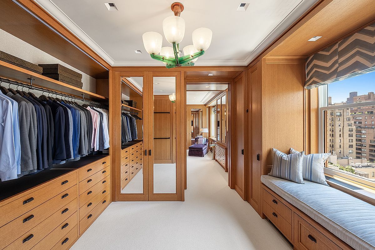 This sophisticated walk-in closet features extensive light wood cabinetry, including hanging rods, drawers, and mirrored doors. A built-in window seat with plush cushions provides a cozy nook, while a unique green-accented chandelier adds a touch of character to the space. The room is finished with neutral carpeting and offers a clear view into an adjacent room, creating a seamless and organized flow.