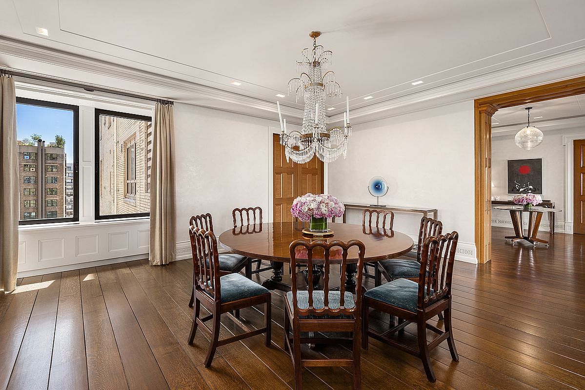 This elegant dining room features a large, round dark wood table surrounded by six upholstered chairs with blue fabric. A grand crystal chandelier serves as the focal point above the table, which is adorned with a vibrant bouquet of pink flowers. The room is framed by tall windows with neutral curtains, rich wood flooring, and a wide doorway leading to an adjacent room with a secondary dining area.