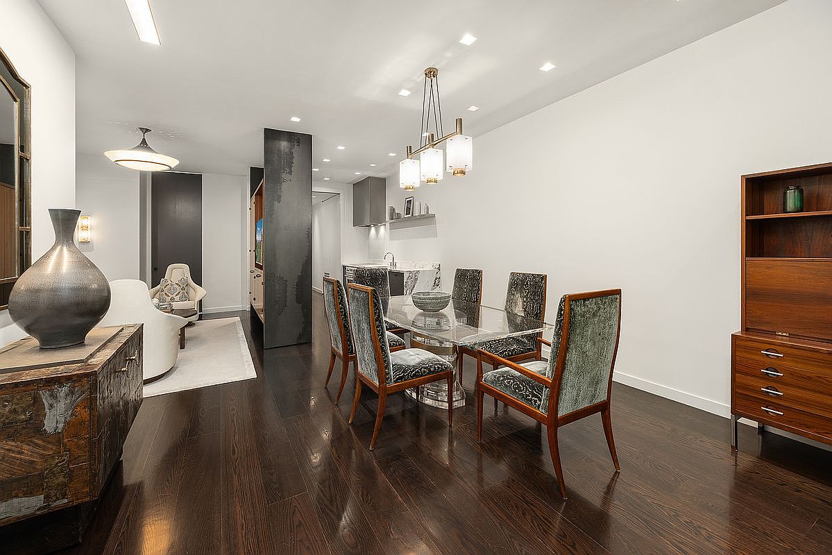 This sophisticated dining area features a glass-topped table surrounded by six chairs upholstered in a textured, patterned fabric. The room is anchored by dark wood flooring and illuminated by a modern, multi-light chandelier, while a tall wooden cabinet provides elegant storage. In the background, a glimpse of a living area and a sleek kitchen creates an open-concept flow, emphasizing a refined and contemporary aesthetic.