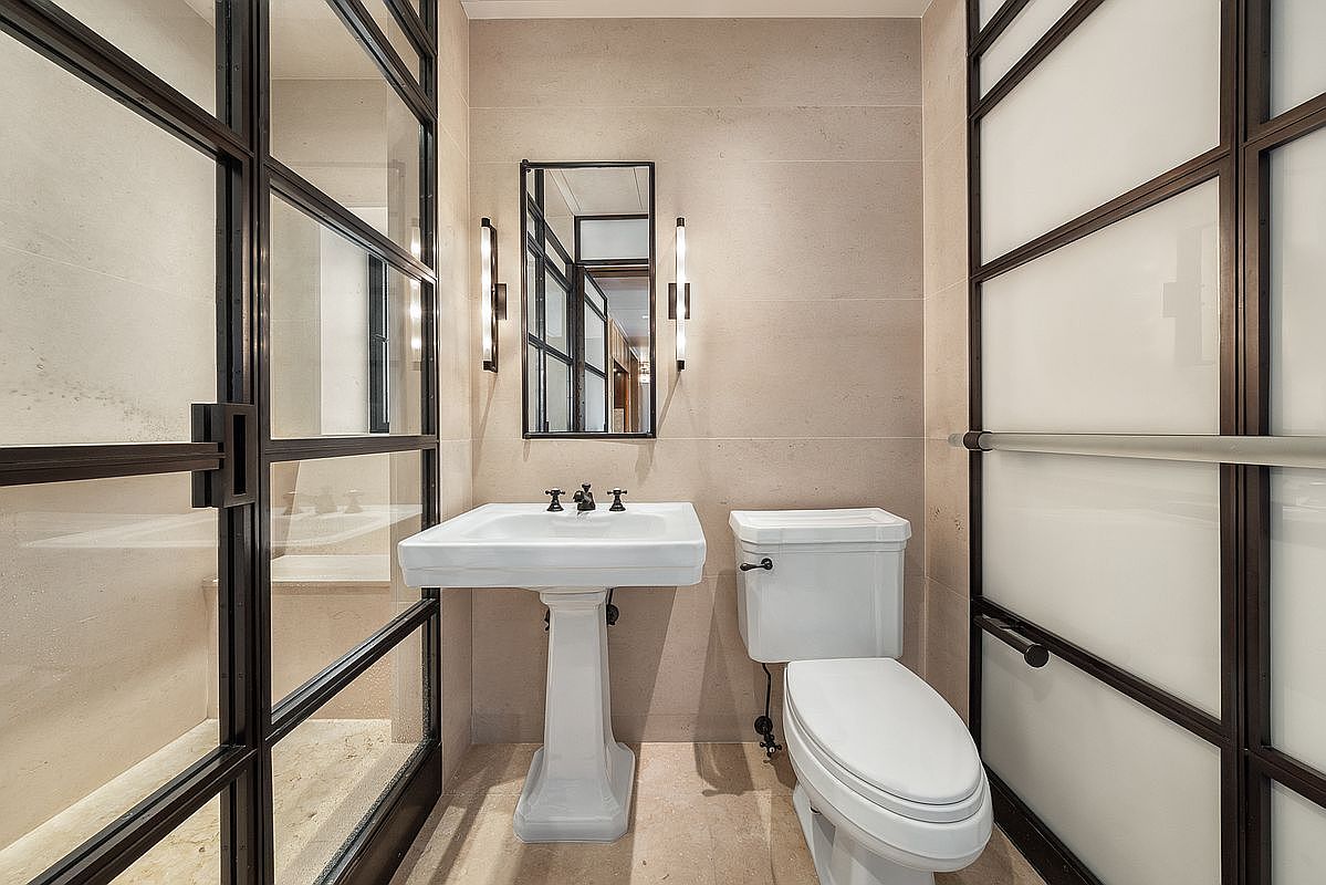 This elegant powder room features a classic pedestal sink and a white toilet set against neutral, large-format wall tiles. The space is framed by striking black-framed glass partitions, including a door on the left and a frosted glass panel on the right, which add a modern, industrial touch. A rectangular mirror is flanked by two vertical wall sconces, creating a balanced and sophisticated aesthetic.