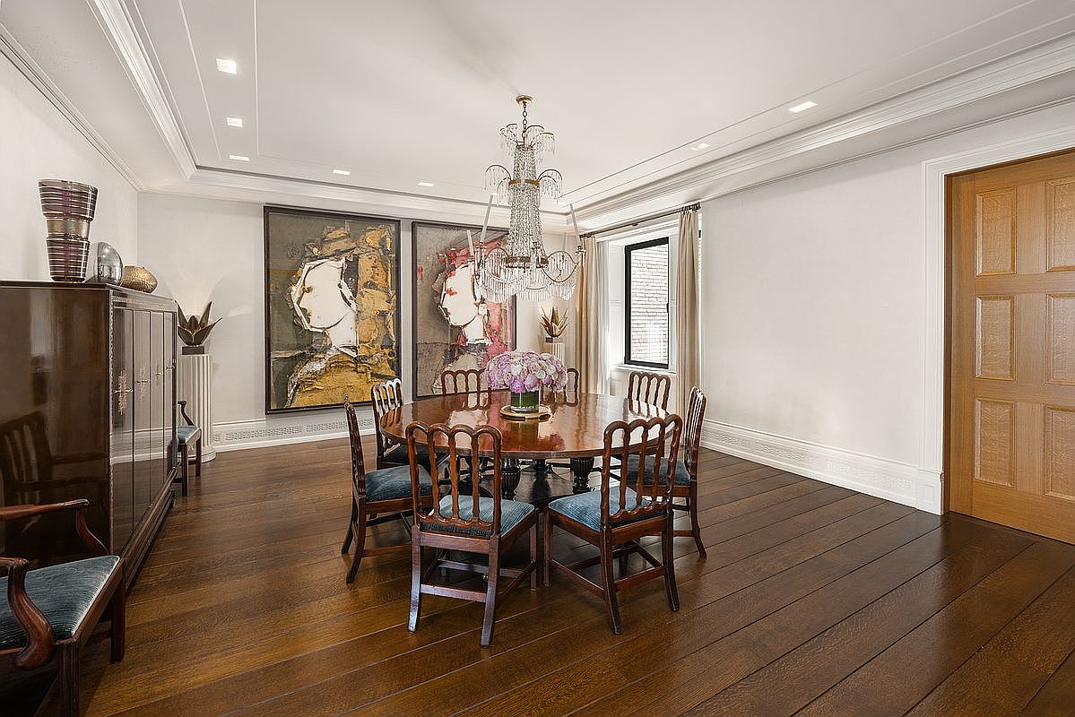 This elegant dining room features dark wood flooring and a large, round wooden table surrounded by six chairs with blue upholstered seats. A crystal chandelier hangs centrally above a floral centerpiece, while two large, abstract portrait paintings serve as a focal point on the wall. The room is framed by white crown molding and a paneled wooden door, creating a sophisticated and formal atmosphere.