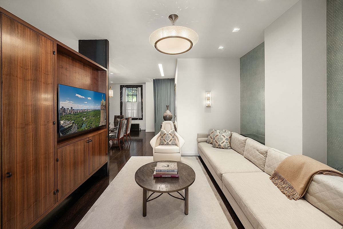 This sophisticated living room features a long, neutral-toned sofa paired with a central round coffee table and a single accent chair. A large wooden media unit with built-in shelving dominates the left wall, while a textured, light-colored wall panel adds visual interest on the right. The space is illuminated by a modern, circular pendant light and recessed ceiling lights, creating a warm and inviting atmosphere that flows into an adjacent dining area.