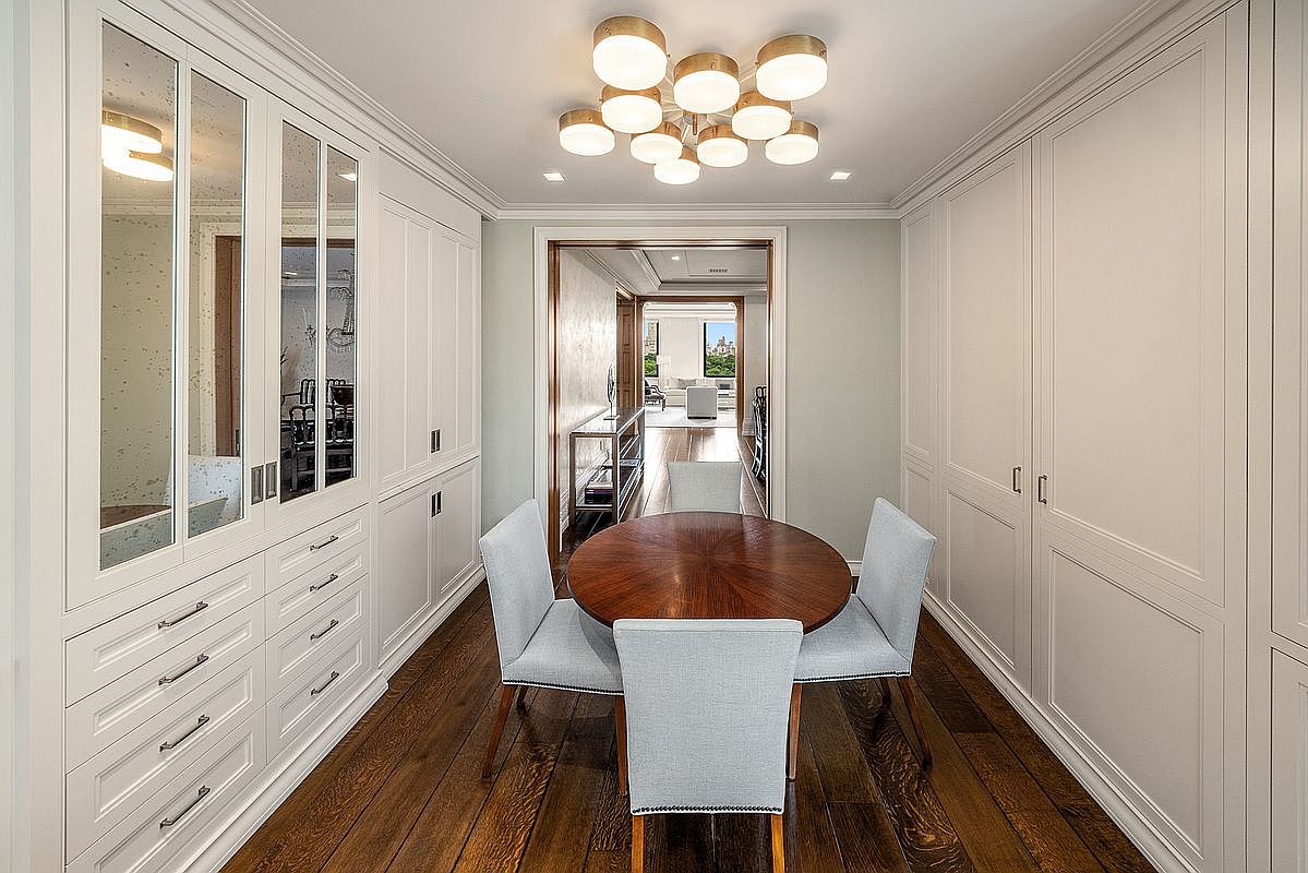 This elegant dining area features a round wooden table surrounded by four light-colored upholstered chairs, set against a backdrop of extensive custom cabinetry with mirrored accents. The room is illuminated by a distinctive multi-globe chandelier and showcases rich dark wood flooring that extends into the adjacent living space visible through the doorway. The overall aesthetic is sophisticated and refined, emphasizing symmetry and high-end built-in storage solutions.