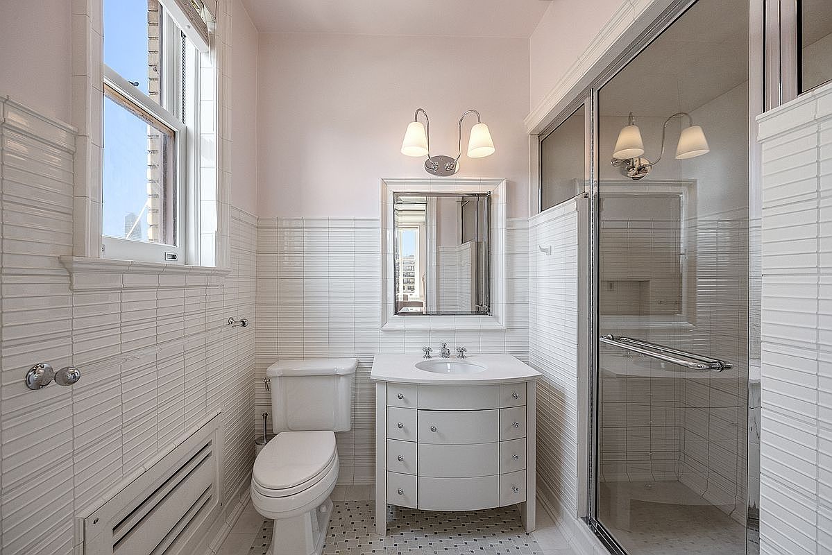 This bright, compact bathroom features white horizontal subway-style wall tiles and a classic mosaic floor pattern. The space includes a white vanity with a stone countertop, a wall-mounted mirror with dual sconce lighting, and a glass-enclosed shower stall. A window to the left provides natural light, creating a clean and well-maintained atmosphere.