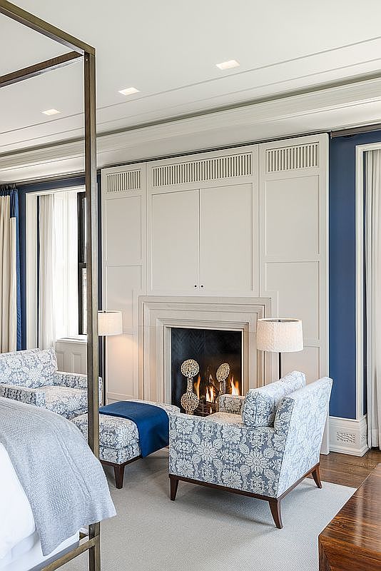 This elegant bedroom features a sophisticated seating area arranged around a central fireplace with a tall, white paneled surround. Two patterned armchairs with a matching ottoman are positioned on a neutral carpet, complemented by a four-poster bed frame in the foreground. The room is accented by deep blue wall panels and tall windows draped in light curtains, creating a refined and tranquil atmosphere.
