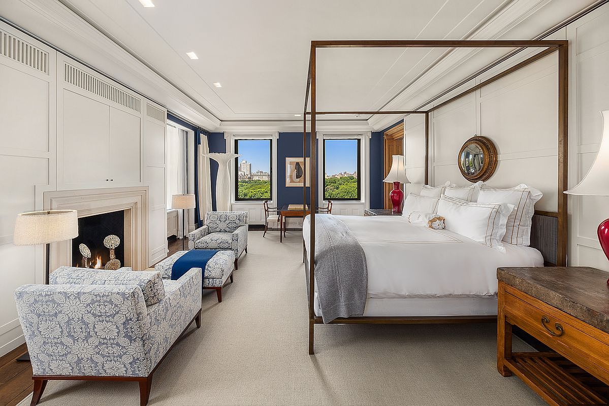 This elegant primary bedroom features a prominent four-poster bed as the central focal point, complemented by a cozy seating area arranged around a fireplace with a classic mantel. The room is decorated in a sophisticated palette of white and deep blue, with large windows that frame expansive views of a lush park. The space is finished with plush carpeting and refined furnishings, creating a luxurious and serene atmosphere.