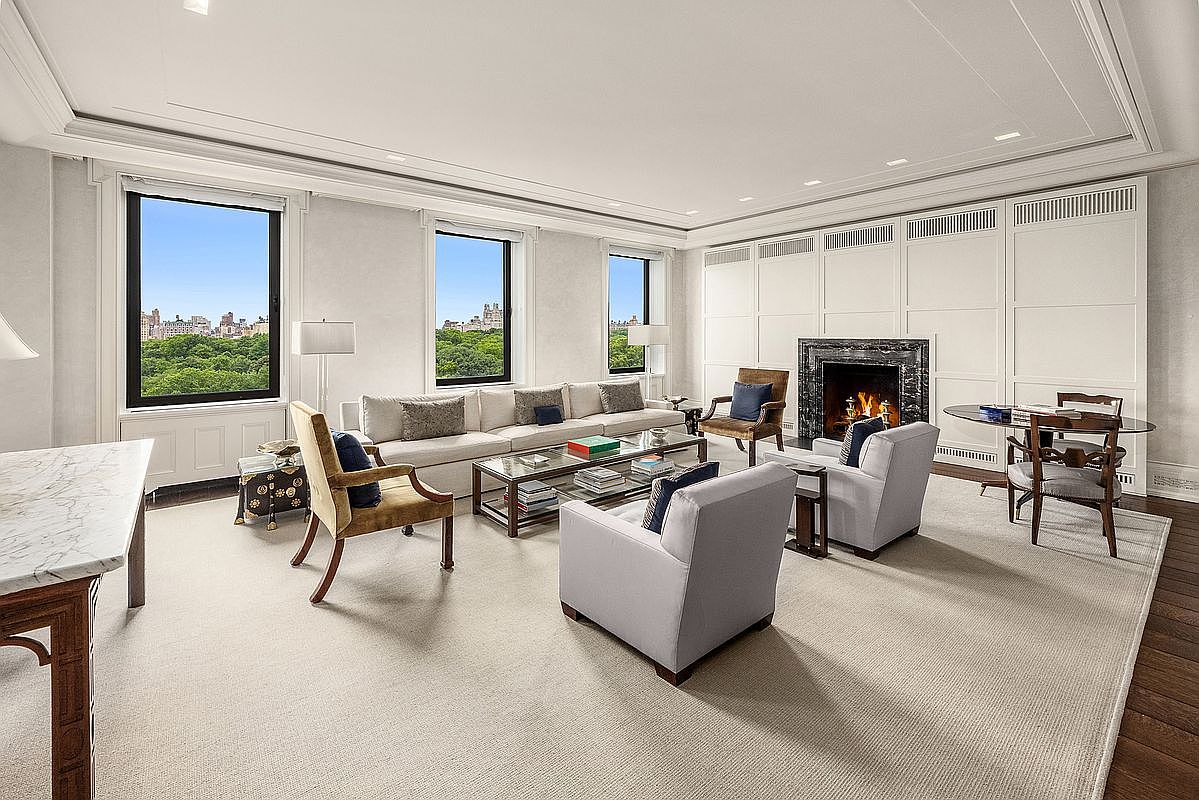 This spacious, light-filled living room features a neutral color palette and a sophisticated, classic design. The room is anchored by a fireplace with a dark stone surround and includes a large sofa, several armchairs, and a glass-topped coffee table arranged on a plush area rug. Large windows provide expansive views of a lush green park, creating a seamless connection between the elegant interior and the natural landscape outside.