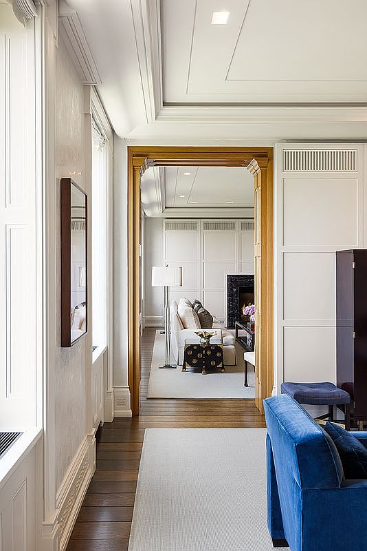This elegant interior view captures a transition space featuring dark wood flooring and crisp white wall paneling. A prominent wooden doorway frames a view into an adjacent living area, which is furnished with a light-colored sofa, a decorative side table, and a dark fireplace surround. The perspective emphasizes the depth and architectural symmetry of the home, highlighting the sophisticated transition between rooms.