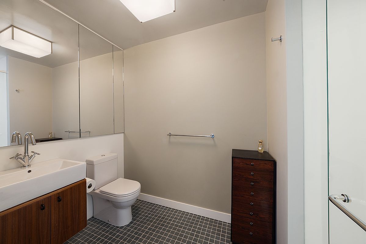 This clean, modern bathroom features a floating vanity with a wood-toned cabinet and a white rectangular sink. A large mirror spans the wall above the vanity, reflecting the space and enhancing the sense of depth. The room is finished with dark, small-format floor tiles and a tall, dark wood chest of drawers, creating a sleek and functional aesthetic.
