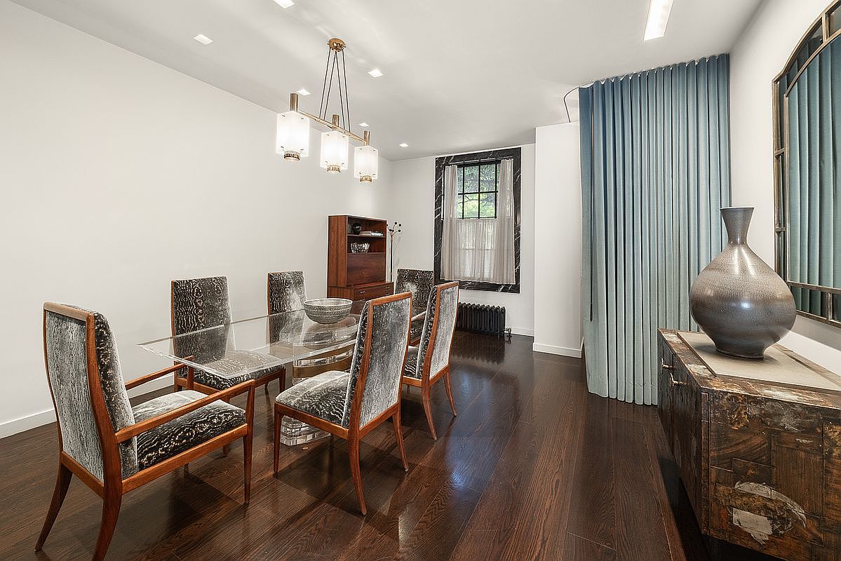 This elegant dining room features a glass-topped table surrounded by six chairs upholstered in a textured, patterned fabric. The space is anchored by dark wood flooring and illuminated by a modern, multi-light chandelier, while a large, dark-framed window and a tall, blue curtain wall add architectural interest. A decorative sideboard with a large, dark vase sits to the right, completing the sophisticated and polished aesthetic of the room.