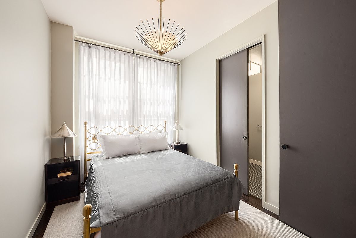 This compact, modern bedroom features a brass-framed bed with a grey coverlet, centered against a window dressed in sheer white curtains. A unique, starburst-style pendant light hangs from the ceiling, adding a touch of mid-century flair to the neutral-toned space. To the right, a dark grey door leads into an adjacent room, while a small nightstand with a metallic lamp sits to the left, creating a clean and minimalist aesthetic.