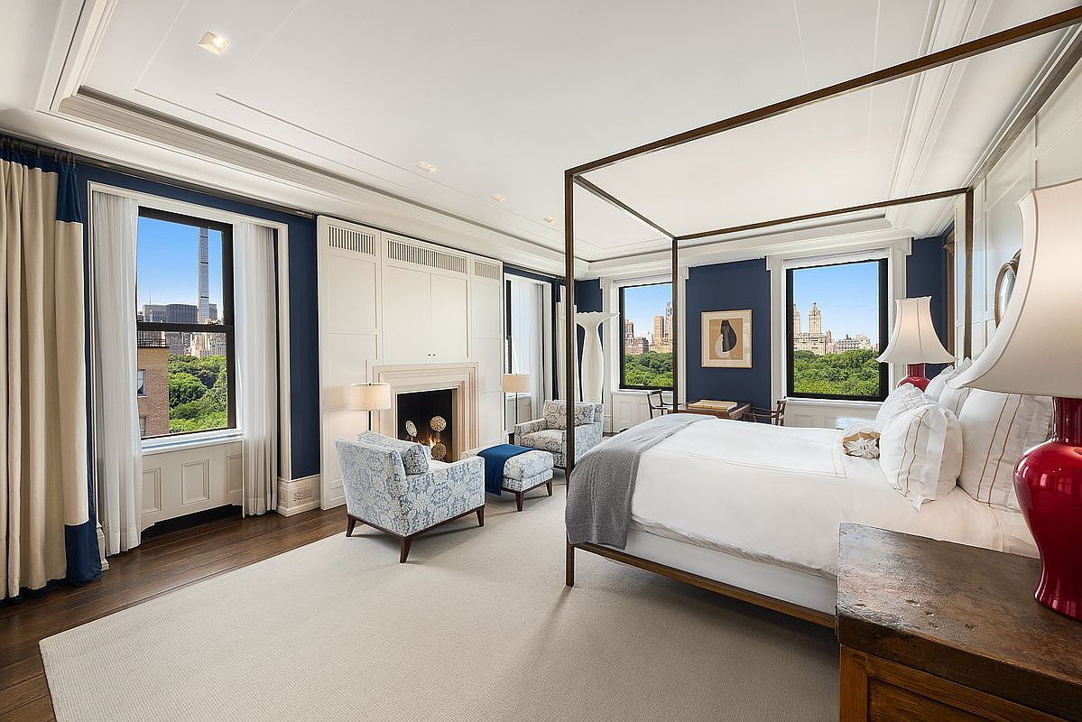 This elegant primary bedroom features a sophisticated navy blue color palette contrasted by crisp white wall paneling and a large area rug. A prominent four-poster bed serves as the centerpiece, complemented by a cozy seating area near a fireplace with a tall, classic surround. Large windows provide expansive, elevated views of a lush green park and city skyline, creating a serene and luxurious atmosphere.