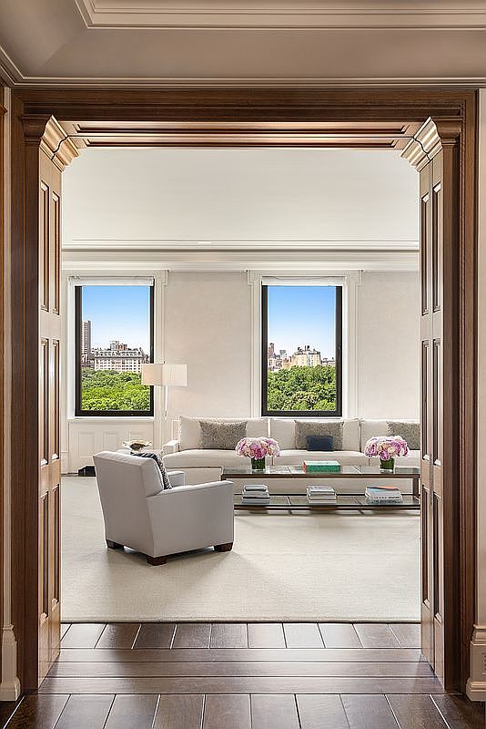 This elegant living room is viewed through a grand, dark-toned wooden doorway, creating a framed perspective of the sophisticated space. The room features a neutral color palette with a plush sofa, a light-colored armchair, and a glass-topped coffee table adorned with floral arrangements. Large windows provide an expansive view of a lush green park and the city skyline beyond, filling the room with natural light.