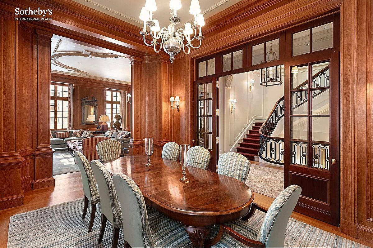 This elegant dining room features rich, dark wood paneling throughout, creating a sophisticated and traditional atmosphere. A large, polished wooden table is surrounded by patterned upholstered chairs, all illuminated by a grand crystal chandelier. The perspective looks through a set of glass-paned double doors into a grand foyer with a sweeping staircase, emphasizing the home's luxurious scale and architectural detail.